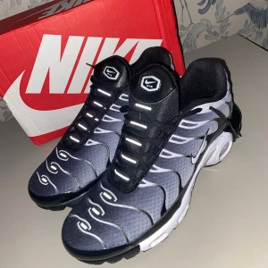 Nike Air Max Plus Tn i svart och grå - Nike Air Max Plus Tn sneakers med svart och grå mesh, vita och svarta vågformade detaljer och reflekterande snörning. Snygg Tn-logga på häl och sula, samt synlig Air Max-dämpning. Perfekta för dig som vill sticka ut med en sportig och modern look.