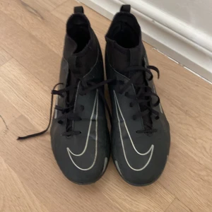 Nike Phantom GT svarta fotbollsskor med bra skick  - Svarta Nike Phantom GT fotbollsskor med vita detaljer och stora Swoosh-loggor. Skorna har snörning, högre strumpliknande skaft och dobbar för bra grepp på planen. Tillverkade i syntetmaterial för lätt känsla och bra bollkontroll.