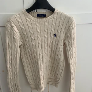 Beige kabelstickad tröja Polo Ralph Lauren - Kabelstickad beige tröja från Polo Ralph Lauren med klassisk rund hals och broderad blå logga på bröstet. Tröjan är i bomull och har långa ärmar samt ribbade. Perfekt för lager-på-lager och snygg till jeans eller kjol. Den har ett litet hål i nacken men syns knappt