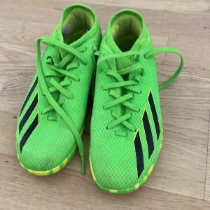 Gröna Adidas fotbollsskor med snörning mycket bra skick  - Snygga fotbollsskor från Adidas i neon-grönt med svarta ränder på sidorna. Skorna har snörning och en mönstrad sula i gult och grönt. Ovandelen är i syntetmaterial med meshstruktur för bra ventilation. Perfekta för dig som vill sticka ut på planen.