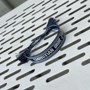 Dior Armband  - Snyggt blått armband från Christian Dior med texten 'CHRISTIAN DIOR J'ADIOR' broderad i vitt. Armbandet är gjort i vävd textil med tvinnade band och tofsar i ändarna. Perfekt accessoar för att lyfta din outfit med en lyxig touch.