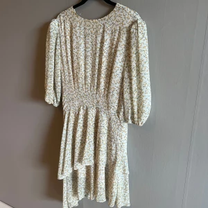 Blommig Adooreklänning  - Supersöt klänning i ljusbeige med små gula blommor. Klänningen har volangig och asymeterisk kjol, trekvartsärmar med lätt puff och markerad midja. Dragkedja på baksidan. Perfekt för vår och sommar! 
