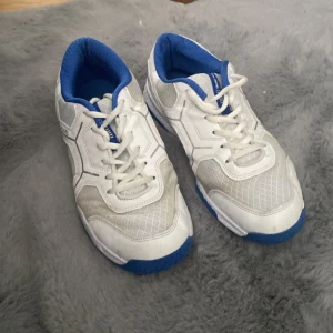 Vita och blå sneakers från Salming - Snygga sneakers från Salming i vitt med blå detaljer och blå sula. Ovandelen är i mesh och syntet för bra andningsförmåga. Skorna har snörning och rund tå, samt Salming-logga och svensk flagga bak. Perfekta för dig som gillar sportig stil.