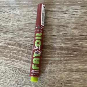 NYX Fat Oil Slick Click Lip Balm - Endast swatchad, NYX Fat Oil Slick Click Shiny Lip Balm i en brunröd nyans. Stiftet har en smidig klickmekanism och ger glansiga, återfuktade läppar. Perfekt för dig som vill ha både färg och glans i ett. Kommer i en praktisk tub med limegröna detaljer.