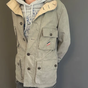 Moncler parkajacka gammal kollektion - Snygg beige parkajacka från Moncler med flera stora fickor framtill och coola knappar. Jackan har en klassisk militärinspirerad look och Moncler-logga på insidan. Perfekt för dig som vill ha en stilren och funktionell jacka till vardags.