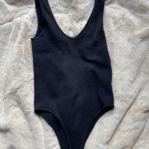 Svart body - Svart ribbad bodysuit med djup rundad urringning både fram och bak. Tight passform och ärmlös design, perfekt att styla med jeans eller kjol för en clean look.