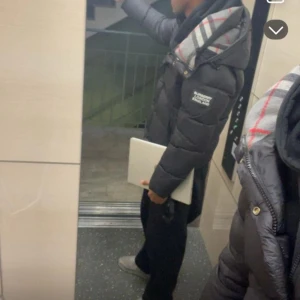 Svart dunjacka från Burberry - Säljer en svart dunjacka från Burberry med klassiskt rutigt foder i huvan. Jackan har lång ärm, dragkedja och knappar framtill samt två fickor med dragkedja. Perfekt för kalla dagar och riktigt snygg streetstil.