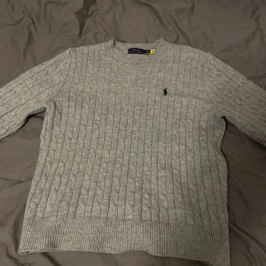 Grå stickad Ralph lauren tröja  - Mycket snygg kabelstickad grå tröja från Ralph Lauren med klassisk rund hals och broderad logga på bröstet. Tröjan är långärmad och har ribbade muddar vid ärmslut och nederkant. Perfekt för dig som gillar stilren och tidlös design.
