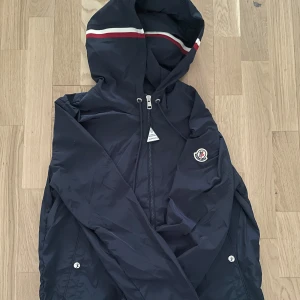 Moncler Windbreaker - Snygg mörkblå vindjacka från Moncler med huva och dragkedja. Jackan har Moncler-logga på armen och vita samt röda ränder på huvan. Klassisk passform med långa ärmar och praktiska fickor med tryckknappar. Vattentät.