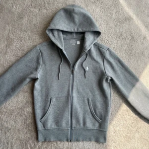 Grå hoodie från Polo Ralph Lauren - Snygg grå hoodie från Polo Ralph Lauren med dragkedja, huva med snörning och klassisk logga broderad på bröstet. Tröjan har två fickor framtill och är tillverkad i mjuk bomullsmix. Perfekt för en chill och stilren look.