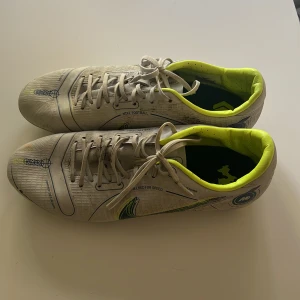 Nike fotbollsskor vit/gul - Snygga fotbollsskor från Nike i vitt med detaljer i neongult och mörkblått. Skorna har snörning, rund tå och platt sula med dobbar för bra grepp på planen. Insidan är fodrad i neongult och ovandelen är i syntetmaterial. Perfekta för dig som vill sticka ut på fotbollsplanen. Behöver bara tvättas sen är dom som nya. 