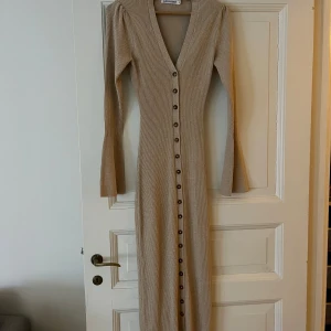 Beige ribbad långklänning från Adoore - Supersnygg beige långklänning från Adoore med ribbad struktur, v-ringning och knappar hela vägen framtill. Klänningen har långa ärmar med lätt utsvängda ärmslut och sitter figurnära för en clean och trendig look.