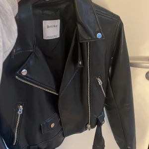 Svart skinnjacka från Bershka - Snygg svart skinnjacka från Bershka i klassisk bikerstil. Jackan har silvriga dragkedjor, knappar och ett bälte i midjan. Perfekt för dig som vill ha en edgy look. Modellen är kortare och har flera fickor med dragkedja.