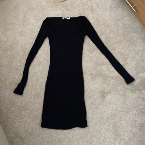 Svart ribbad klänning från NA-KD - Snygg svart ribbad klänning från NA-KD med lång ärm och fyrkantig ringning. Modellen är tight och kort, perfekt för dig som gillar en stilren look. Materialet är stretchigt och sitter skönt på kroppen.