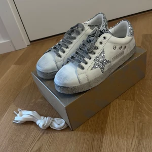 Golden Goose Superstar sneakers glitter - Golden Goose Superstar sneakers i vitt skinn med silverglittrig stjärna och häl. Grå snörning med logga, rund tå och slitna detaljer på sulan för en cool look. Extra vita skosnören medföljer. Perfekta för dig som gillar statement-sneakers. Helt ny oanvänd 