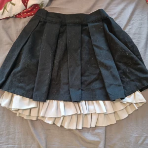 Svart plisserad kjol med vit volang - Supersöt svart kortkjol med breda plisserade veck och en vit volangkant som sticker fram nedtill. Kjolen har en klassisk skoluniform-vibe och är perfekt för dig som gillar en lekfull och trendig stil. Den går att reglera i midjan. 