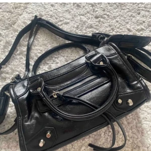 Svart axelväska i skinn med dragkedja - Snygg svart axelväska i skinn med coola silverdetaljer och dragkedja framtill. Väskan har både handtag och lång axelrem, perfekt för att bära på olika sätt. Stilren design med nitar och dekorativa sömmar som ger en edgy vibe.