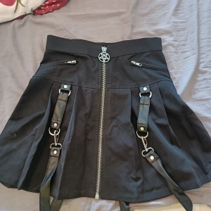 Svart kortkjol med straps från Killstar - Svart kortkjol från Killstar med dekorativa dragkedjor och en stor pentagram-dragkedja framtill. Kjolen har coola straps och metalldetaljer som ger en edgy vibe. Perfekt för dig som gillar alternativ stil.