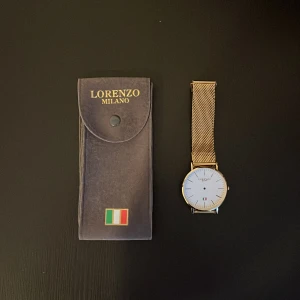 Guldig mesh-klocka Lorenzo Milano - Stilren klocka från Lorenzo Milano med vit urtavla och italiensk flagga. Boetten är rund och armbandet är i guldig mesh, vilket ger en modern och elegant look. Klockan har minimalistisk design med tunna svarta markeringar och justerbart spänne.