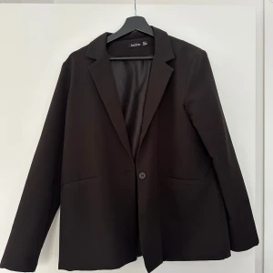 Svart kavaj från Boohoo - Stilren svart kavaj från Boohoo med klassisk krage och enkel knäppning framtill. Kavajen har två fickor och är fodrad för extra komfort. Perfekt för dig som vill ha en tidlös och snygg look.