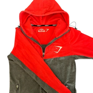 Gymshark röd och grå hoodiejacka M - Snygge hoodiejacka från Gymshark i rött och mörkgrått med dragkedja och huva. Jackan har Gymshark-logga på bröstet och praktiska fickor med dragkedja. Perfekt för träning eller chill, med sportig vibe och bekväm passform.