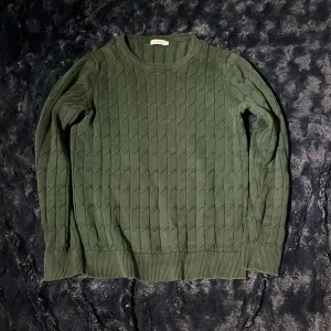 Grön stickad tröja J.Lindeberg - Mörkgrön stickad tröja från J.Lindeberg med snyggt strukturmönster. Tröjan har rund hals, ribbade muddar och lång ärm. Perfekt för dig som gillar stilrena och bekväma plagg med lite edge.