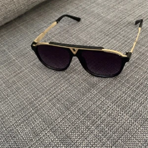 Svarta och guldiga LV solglasögon - Snygga solglasögon med svart båge och guldiga detaljer. Glasen är mörka och har en klassisk aviator-inspirerad form. Perfekta för att lyfta din stil med en lyxig touch. Pris kan diskuteras 
