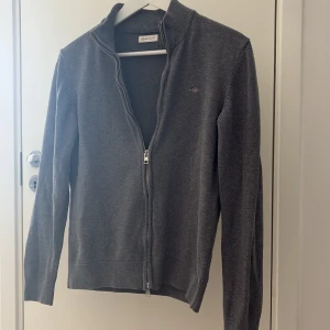Grå zip up tröja Gant - Mörkgrå kofta från Gant med hel dragkedja och diskret broderad logga på bröstet. Tröjan är ribbad vid krage, ärmslut och nederkant. Perfekt för lager-på-lager och passar till det mesta. Tillverkad i mjuk bomull för en skön känsla. Säljer för 400kr ordinarie pris 1200kr. Skick 9/10 använd ett fåtal gånger