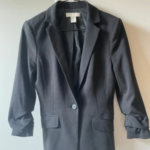 Svart kavaj från H&M, storlek 34 - Klassisk svart kavaj från H&M i storlek 34. Enkelknäppt med två fickor framtill och snyggt skräddad passform. Perfekt för en stilren look. Materialet är mjukt och har en lätt stretch för extra komfort.