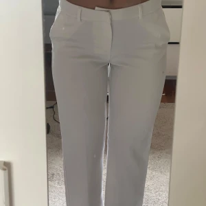 Vita kostymbyxor med raka ben - Säljer ett par stilrena vita kostymbyxor med raka ben och hög midja. Byxorna har hällor för bälte, fickor fram och dekorfickor bak. Perfekta för en clean och fräsch look. Materialet känns mjukt och har en lätt stretch. 