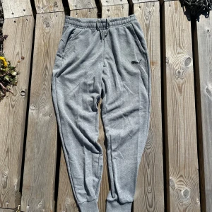 Grå mjukisbyxor från Puma - Snygga grå mjukisbyxor från Puma med elastisk midja och dragsko. Byxorna har ribbade muddar vid bensluten, två sidofickor och Puma-logga på låret. Perfekta för chill eller träning, tillverkade i mjuk bomull. Mått: Waist: Reglerbar. Inside leg length: 78cm 