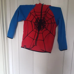 Spiderman fleecehoodie från H&M - Cool röd och blå fleecehoodie från H&M med Spiderman-tryck och broderade ögon på huvan. Tröjan har lång ärm, dragkedja framtill och är perfekt för dig som älskar Marvel. Mjuk och skön fleece som håller dig varm under kyliga dagar.