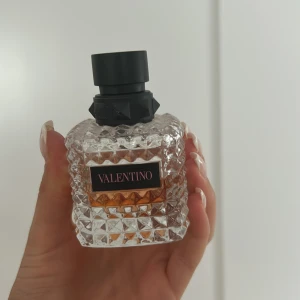 Valentino parfym med nitar - Lyxig parfym från Valentino i en transparent, fyrkantig glasflaska med coolt nitat mönster och svart etikett. Flaskan har en stor svart kork med nitar och innehållet är ljusrosa. Perfekt för dig som gillar exklusiva och trendiga dofter.