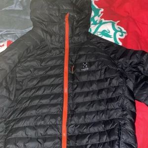 Svart dunjacka från Haglöfs med orange dragkedja - Säljer en svart dunjacka från Haglöfs med snygg orange dragkedja och huva. Jackan är quiltad och har en lätt, sportig känsla. Perfekt för kyliga dagar och outdoor-äventyr. Klassisk Haglöfs-logga på bröstet och praktiska fickor.