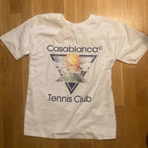 Vit Casablanca Tennis Club t-shirt - Vit t-shirt från Casablanca med tryck framtill som visar sol, moln och texten 'Tennis Club'. Klassisk passform med rund hals och korta ärmar. Perfekt för dig som gillar sportig och stilren vibe.