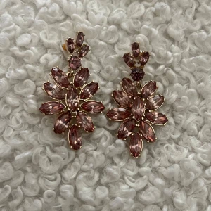 Rosa örhängen - Säljer ett par stora statement örhängen med glittrande rosa stenar i blomform. Örhängena har guldfärgade detaljer och är perfekta för att sticka ut. De är formade som blommor med flera avlånga stenar som ger en lyxig och trendig känsla.