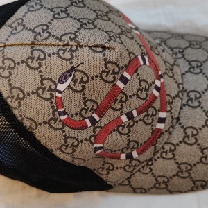 Gucci keps med orm och GG-mönster - Snygg keps från Gucci med klassiskt GG-monogram i beige och svart, prydd med en röd, vit och svart orm framtill. Baksidan är i svart mesh för extra ventilation. Justerbar passform och ikonisk streetstyle-vibe. Knappt använd. 