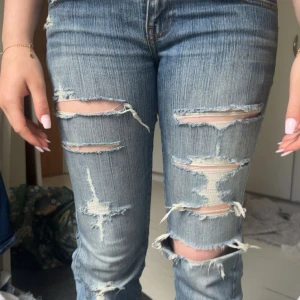 Blå slitna bootcut jeans med hål - Säljer ett par blå bootcut jeans med flera slitningar och hål framtill för en riktigt edgy vibe. Jeansen har klassisk femficksdesign, normal passform och är tillverkade i jeansmaterial. Perfekta för dig som gillar en avslappnad och trendig look. litet hål på insidan av låret men syns inte mycket 