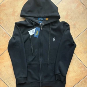Svart hoodie från Polo Ralph Lauren - Svart hoodie från Polo Ralph Lauren med klassisk vit broderad logga på bröstet. Tröjan har huva med snörning, dragkedja framtill och två fickor. Tillverkad i mjuk bomull, perfekt för en chill och stilren look.