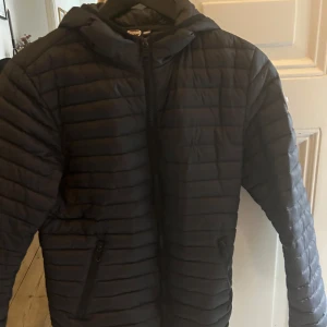 Svart pufferjacka från Colmar - Säljer en svart pufferjacka från Colmar med huva och dragkedja framtill. Jackan har horisontella sömmar, två fickor med dragkedja och en snygg logga på ärmen. Perfekt för kyliga dagar när du vill hålla stilen.