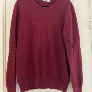 Vinröd tröja från J.Lindeberg strl M - Säljer en stilren vinröd tröja från J.Lindeberg. Tröjan har en subtil textur och rund halsringning. Perfekt för att hålla stilen i kyligare väder. Passar både till jeans och chinos för en avslappnad look. Vinröd stickad tröja i strl M från J.Lindeberg, modell Dexter circle structure.  Använd men i fint begagnat skick.  Mått Över bröstet: ca 53 cm Längd från axeln och ner: ca 68 cm  100% bomull  Kommer från ett djur och rökfritt hem.