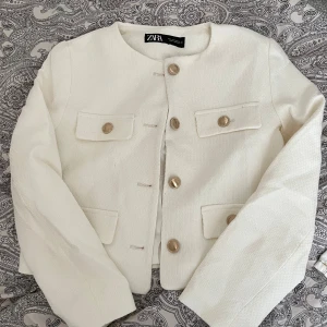 Vit kavaj från Zara - Säljer en stilren vit kavaj från Zara med guldknappar framtill. Kavajen har en klassisk design med rund hals och långa ärmar. Perfekt för en elegant look.