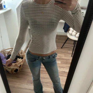 Randig långärmad topp - Säljer en stilren randig topp med långa ärmar. Toppen har en tight passform och är perfekt för både vardag och speciella tillfällen. Den är i en klassisk färgkombination av beige och svart.