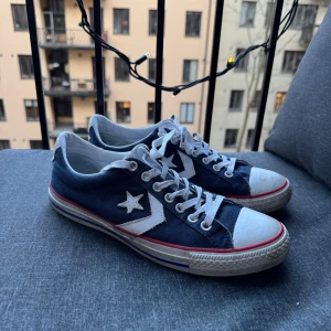  Converse skor - Säljer ett par klassiska blå Converse sneakers med vita detaljer och snörning. Skorna har en vit sula med röda ränder och den ikoniska stjärnloggan på sidan. Perfekta för en avslappnad stil.