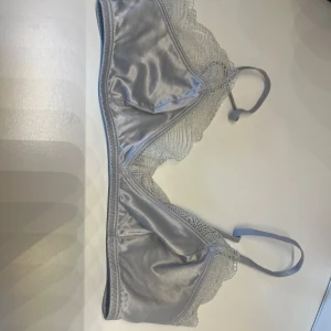 Grå spetsdetaljerad bralette - Snygg grå bralette med spetsdetaljer och justerbara axelband. Perfekt för en stilren och bekväm look. Den har en mjuk och glansig finish som ger en lyxig känsla.