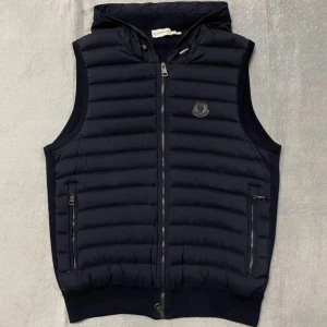 Svart dunväst från Moncler - Snygg svart dunväst från Moncler med dragkedja och huva. Västen har en quiltad design och två praktiska fickor med dragkedja framtill. Perfekt för lager-på-lager under kyligare dagar.