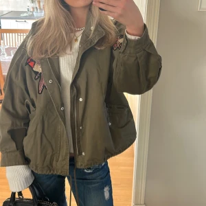 Olivgrön jacka med broderade detaljer - Snygg olivgrön jacka med broderade detaljer på axlarna. Jackan har en dragkedja och knappar framtill samt en loose passform. Perfekt för höst och vår.