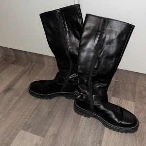 Svarta stövlar från Gabor - Snygga svarta stövlar från Gabor i skinn med dragkedja på sidan. De har en robust sula och en dekorativ remdetalj runt ankeln. Perfekta för både stil och komfort.