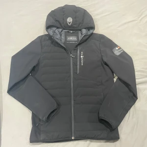 Svart jacka från Sail Racing - Snygg svart jacka från Sail Racing med huva och dragkedja. Jackan har en cool patch på ärmen med texten 'Sail Antarctica Expedition'. Perfekt för kyliga dagar med sin quiltade design och långa ärmar. Pris är ej hugg i sten!
