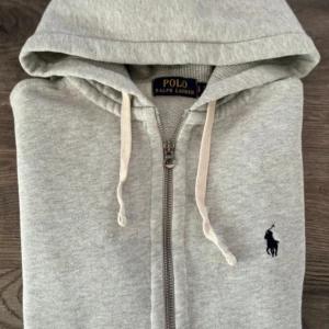 Grå hoodie från Ralph Lauren - Säljer en stilren grå hoodie från Ralph Lauren med dragkedja och justerbar huva. Tröjan har en broderad logotyp på bröstet och praktiska fickor framtill. Perfekt för en avslappnad look.